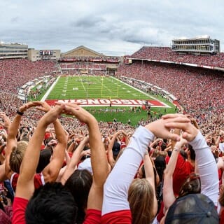 Fans hold their arms up to make an "O" with the football field in the background.