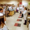 a group of people in white lab coats stand in a circle in a biotechnology lab listening to a speaker.