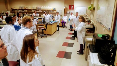 a group of people in white lab coats stand in a circle in a biotechnology lab listening to a speaker.