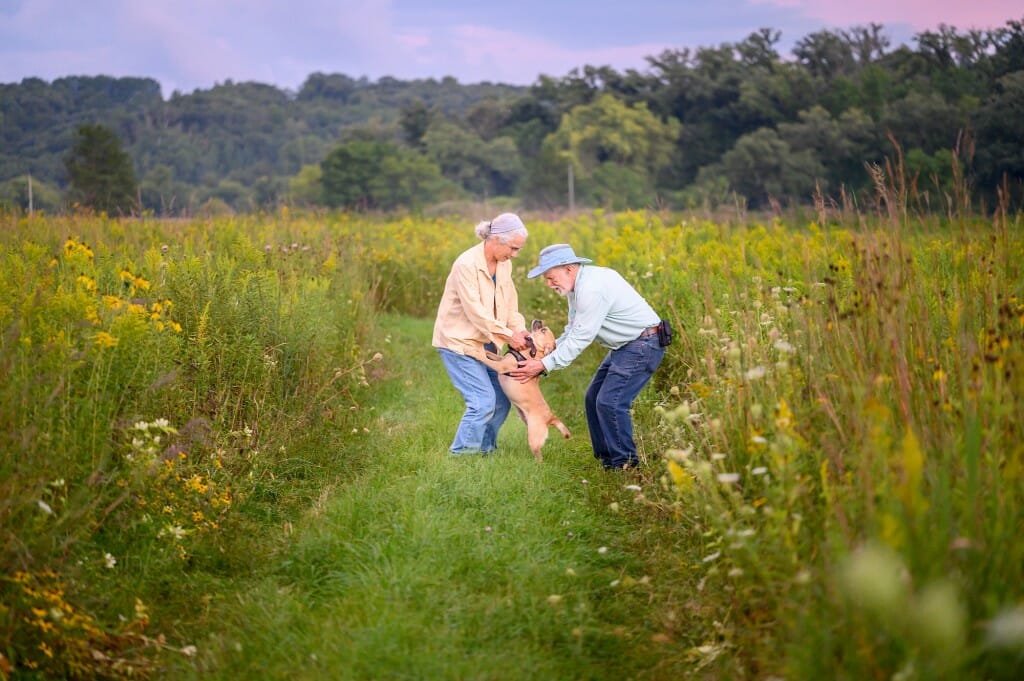 A woman and a man stand in a prairie petting a small dog