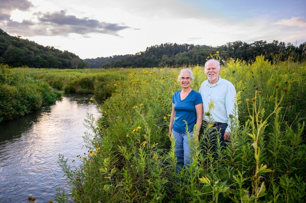 A couple stands in a meadow next to a stream.