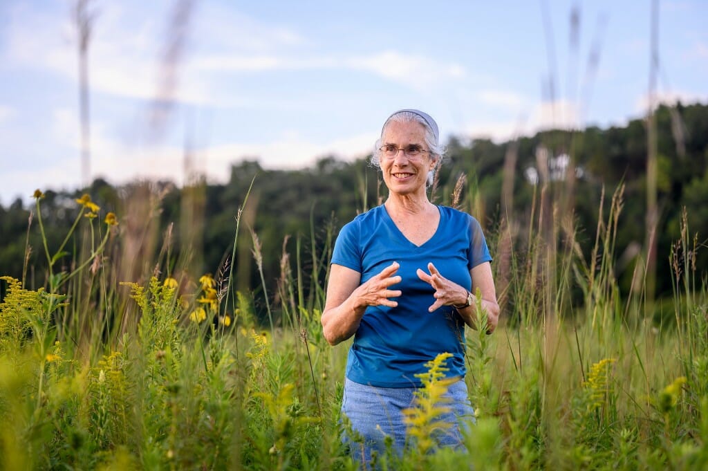 A woman in a blue shirt stands in a meadow and gestures with her hands.