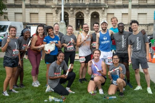 A group of runners stand on a lawn and hold up their "diplomas" received for finishing the race.