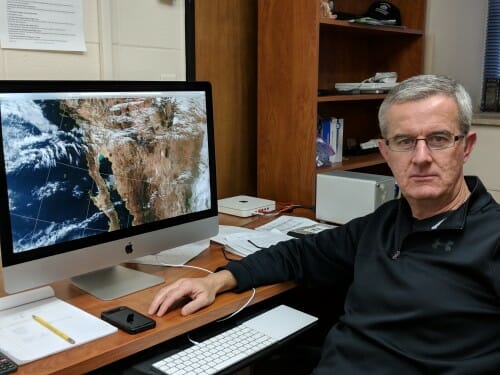 Liam Gumley sits at a desk in front of a computer monitor showing a satellite image of North America