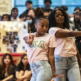 Two dancers wearing pink Alpha Kappa Alpha sorority T-shirts, pearls and light-wash jeans move in unison with their left arms outstretched and pinky fingers raised.