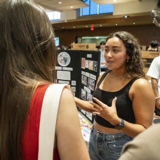 A close shot of a student wearing a black top and jeans stands at an exhibit booth speaking to two students visiting her booth.