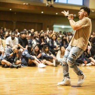 A man wearing a tan T-shirt, faded jeans and white sneakers sings into a microphone in front of a seated audience.