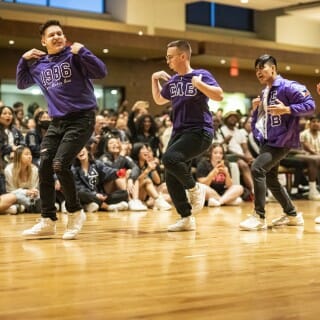 Four dancers in purple Sigma Lambda Beta fraternity insignia dance in line. A seated audience cheers in the background.