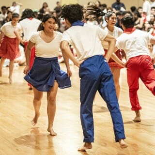Dance couples wearing red, blue and white dance barefoot on a wooden floor. A seated audience watches in the background.