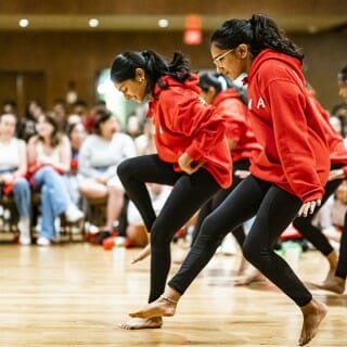 Five women in red hoodies and black leggings dance barefoot on a wooden floor. A seated audience watches in the background.
