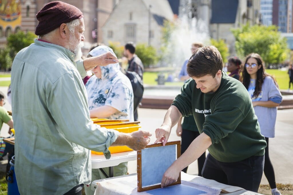 Robert Russehl gestures with one hand while a student begins to pull wet paper from a paper making screen.