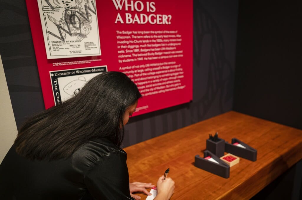A woman writes on a paper in front of a table, with an exhibit in the background.