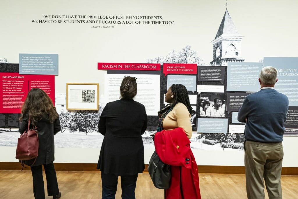 Four visitors stand facing away from the camera to view an exhibit wall with a banner reading "Racism in the Classroom."