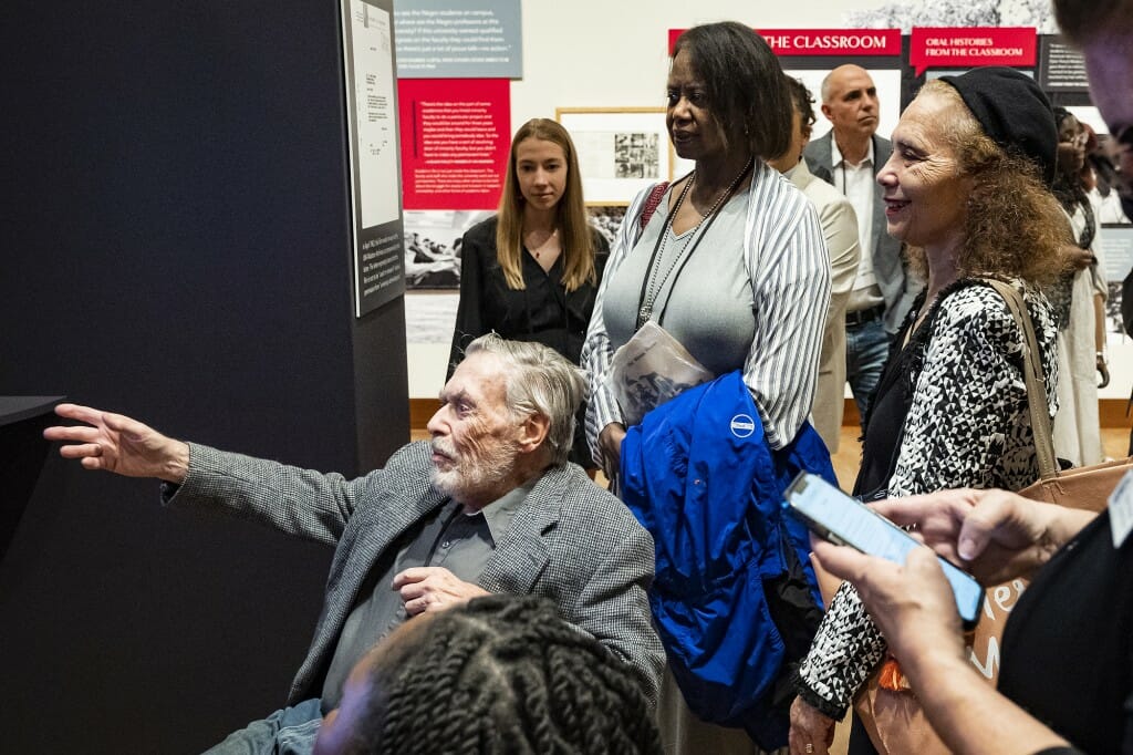 A small crowd stand around a seated man who gestures with an open hand toward an unseen wall exhibit.