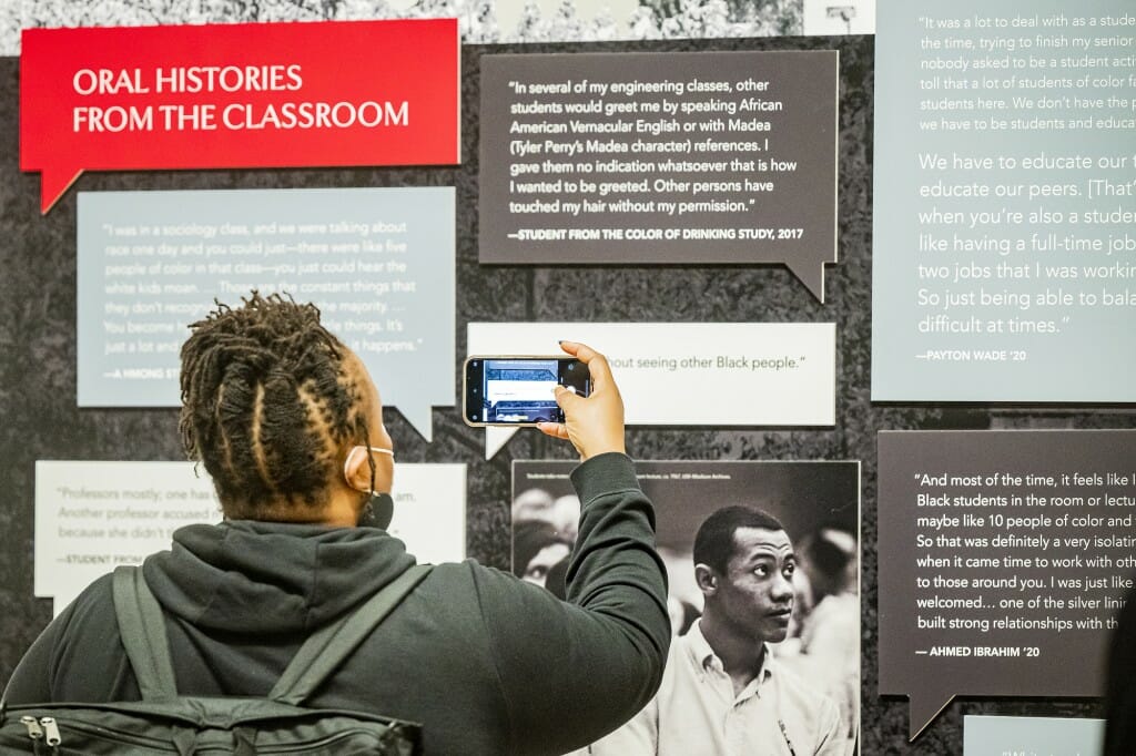 An attendee faces away from the camera toward an exhibit wall. They raise their phone to take a photo of an exhibit wall with the banner "Oral histories from the classroom."