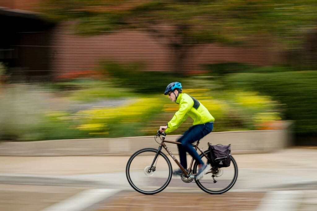 A cyclist wearing a neon jacket and black pants bikes in light rain past a flowerbed and a red brick building