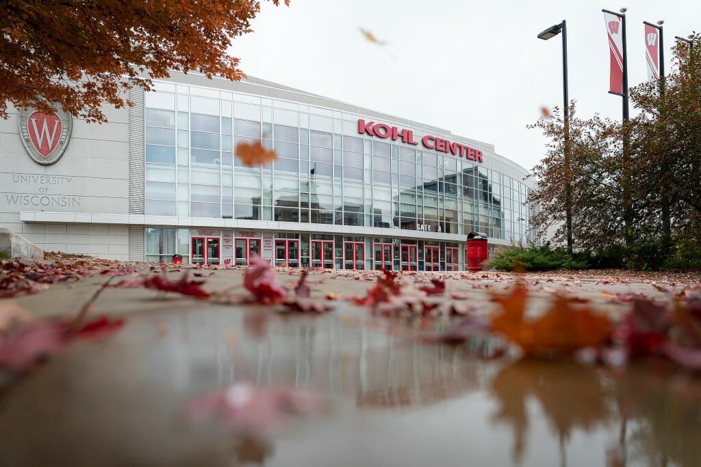 Low perspective looking up at the Kohl Center. Red maple leaves cover the wet sidewalk in front of the building's windowed facade.