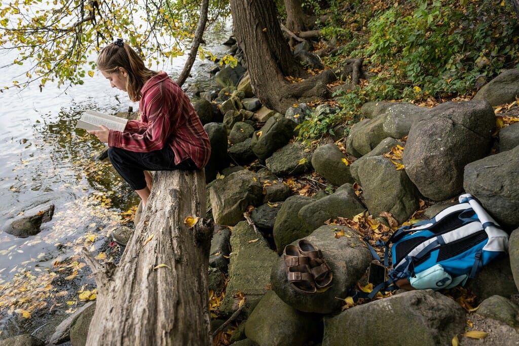 Undergraduate student Sofie Schachter sits on a log on the shore of Lake Mendota reading a book.