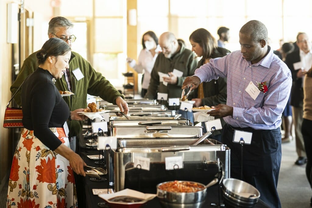 People serve themselves from a buffet line in a brightly sunlit room.