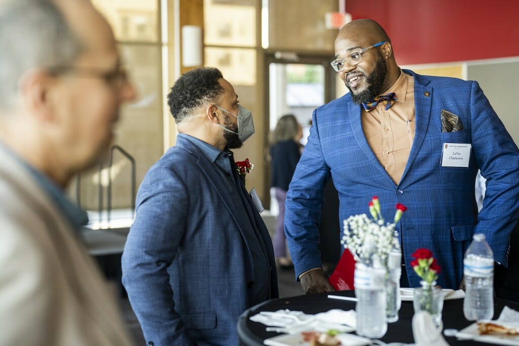 LaVar Charleston (right) speaks with Reginald Royston (center). Both wear blue blazers and stand behind a cocktail table in a brightly sunlit room.