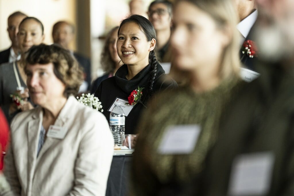 Jessica Hua smiles and looks ahead in a crowd of people listening to remarks from Chancellor Mnookin.