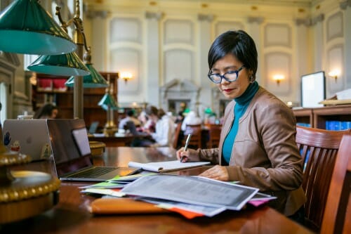Monica Kim sits at a table in the reading room of the Wisconsin Historical Society. She is taking notes while reading from a stack of papers.