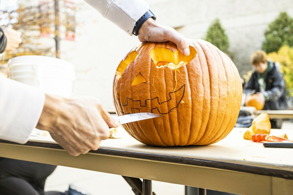 A pumpkin has eyes and nose carved already, and a knife is poised to cut out the mouth.