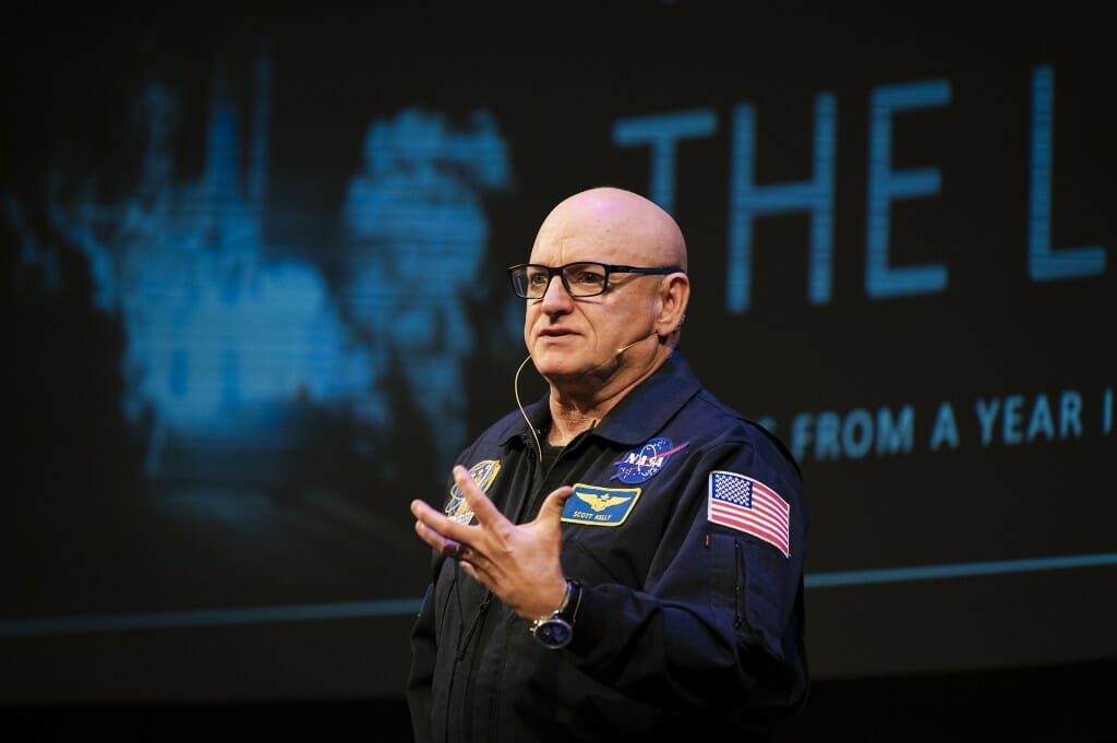 A close shot of Scott Kelly wearing his NASA flight suit in front of a dark stage background.