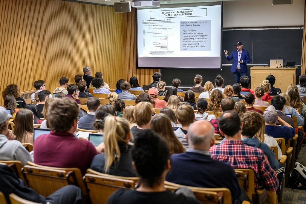 Nate Silver gives a lecture in a Russel Laboratories classroom. He stands at the front of the class and gestures to a projector screen showing slides from his presentation.