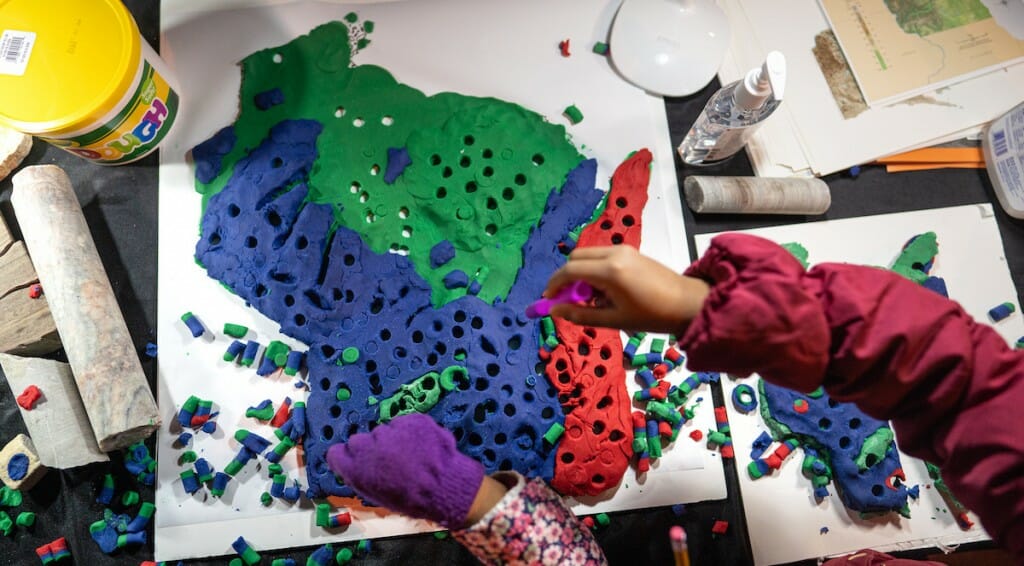 An overhead view of a table top covered in craft supplies. In the center, childrens' hands reach over a playdough sculpture of the state of Wisconsin.