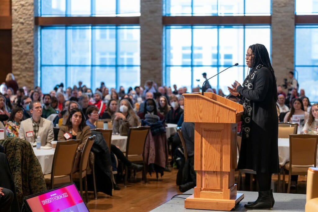 A woman speaks at a podium to a large crowd, with large windows backlighting the scene.