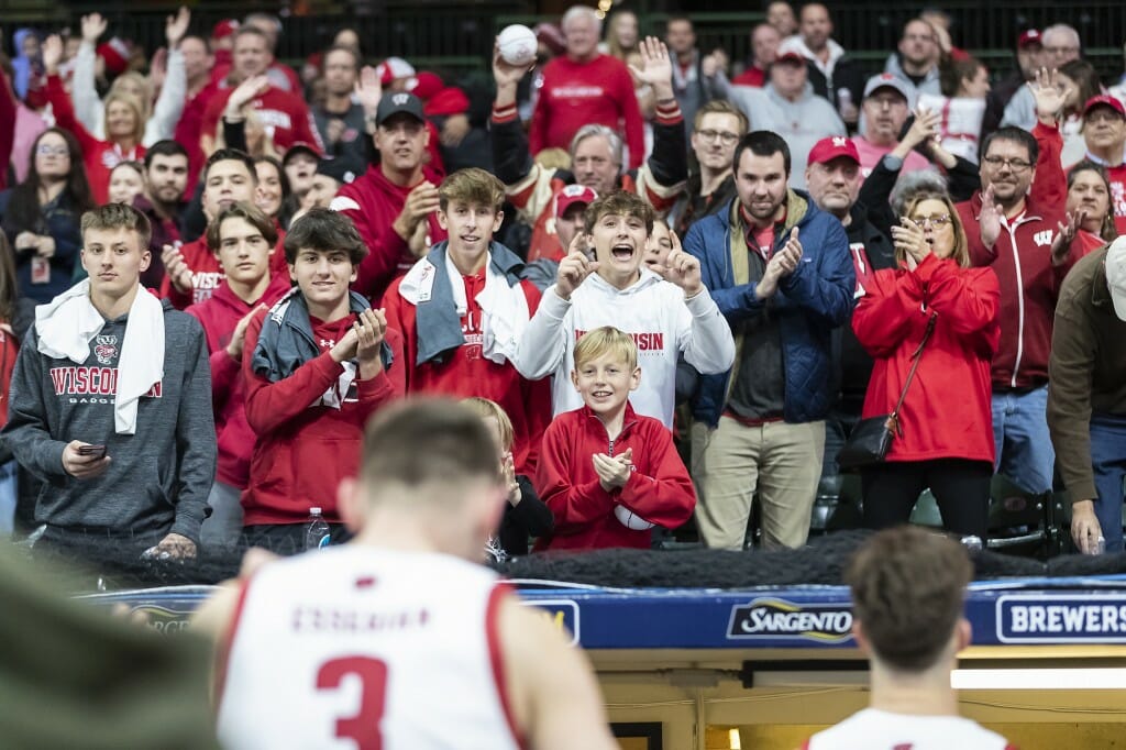 People in stands stand, wave their arms and shout. A Wisconsin basketball player stands in the foreground.