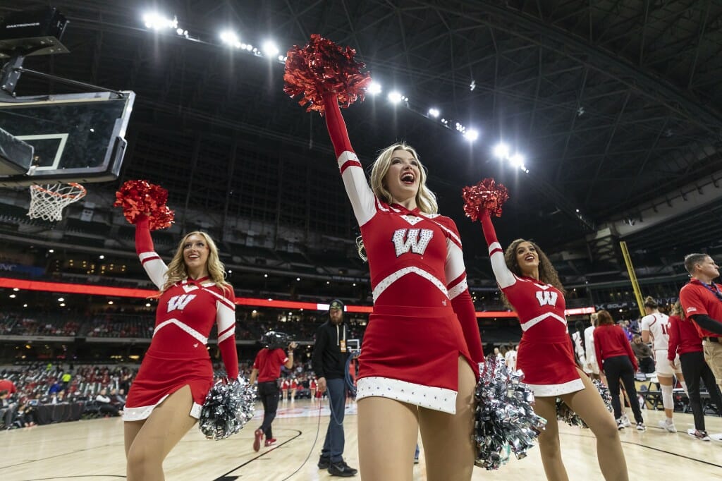 A group of cheerleaders stand on the court, raise their arms and shout.