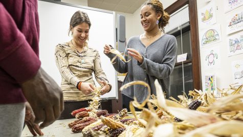 Two women laugh and talk as they braid corn, with a pile of corn cobs in the foreground.