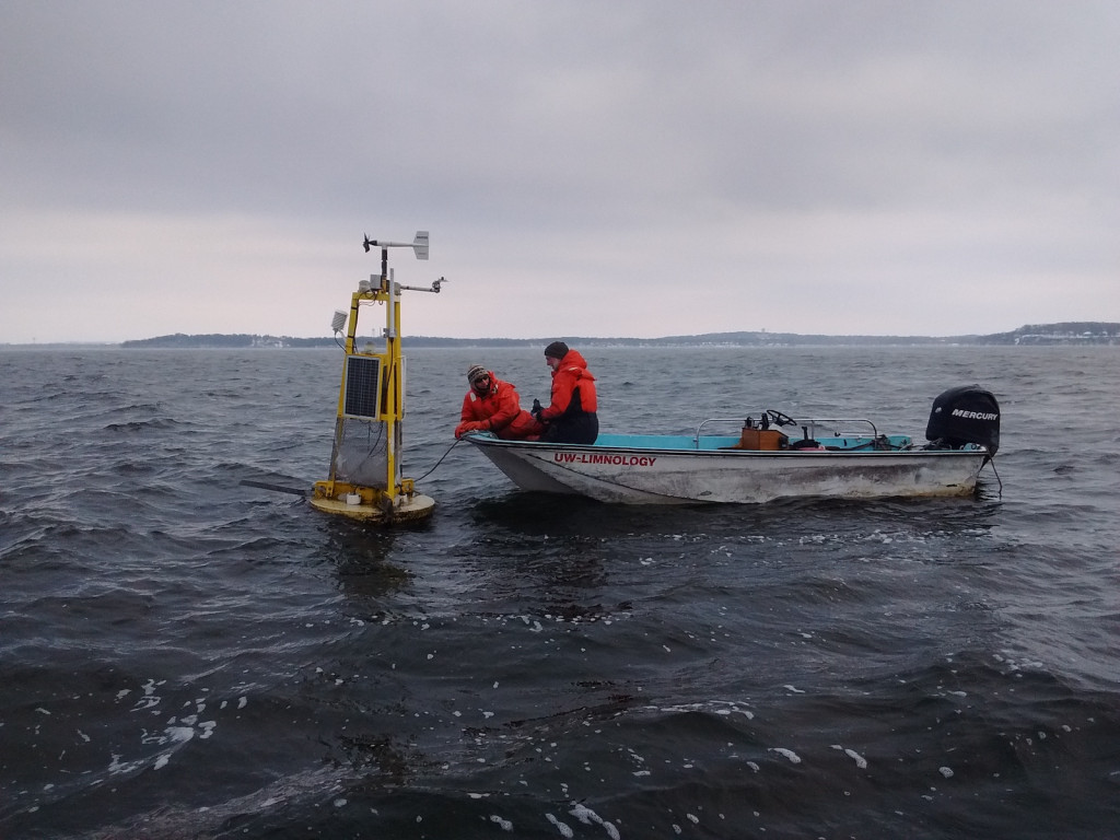 Two men are in a boat on a gray day on Lake Mendota. They lean over to take readings from a buoy.