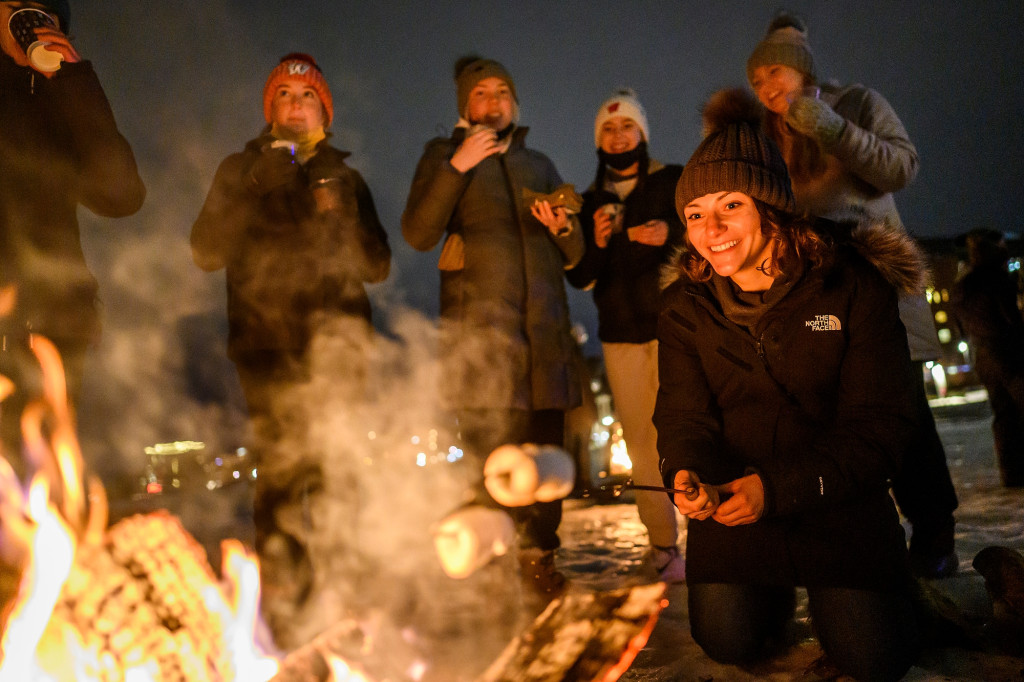 Students stand around a bonfire at night on Lake Mendota. One student crouches down and holds roasting sticks stacked with marshmallows over the open flame.