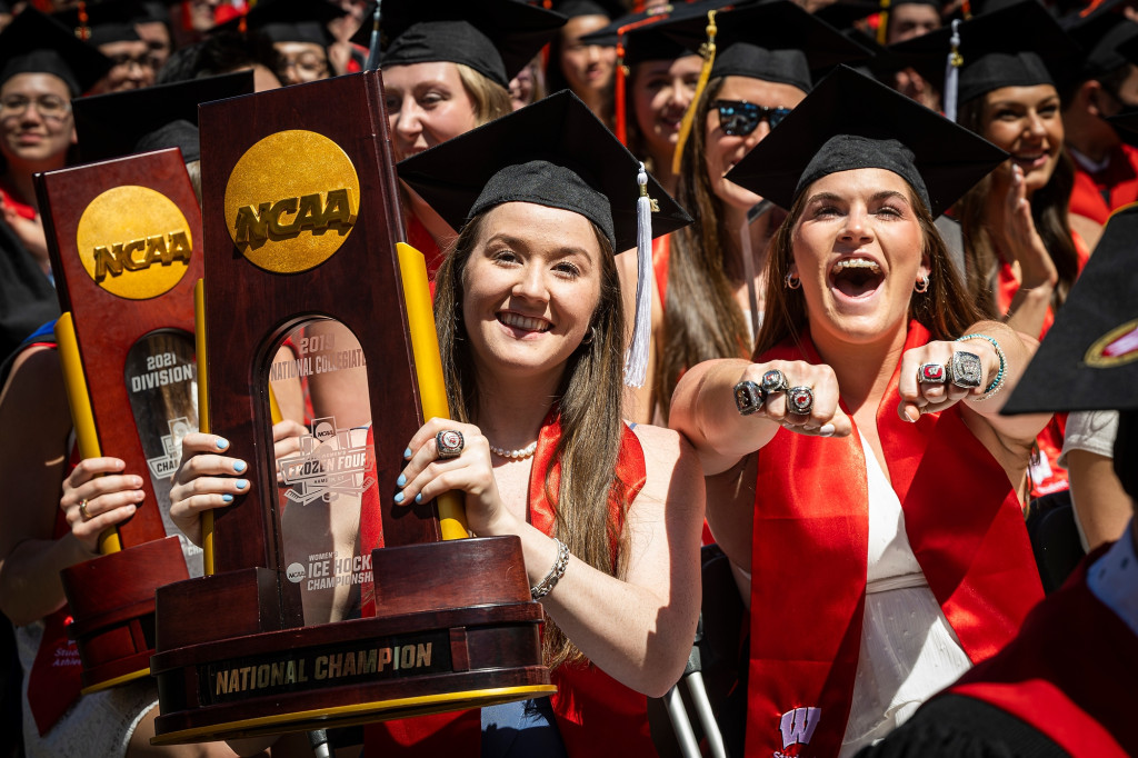 Members of the UW women’s volleyball and hockey teams hold their trophies and smile toward the camera while wearing caps and gowns at spring commencement.