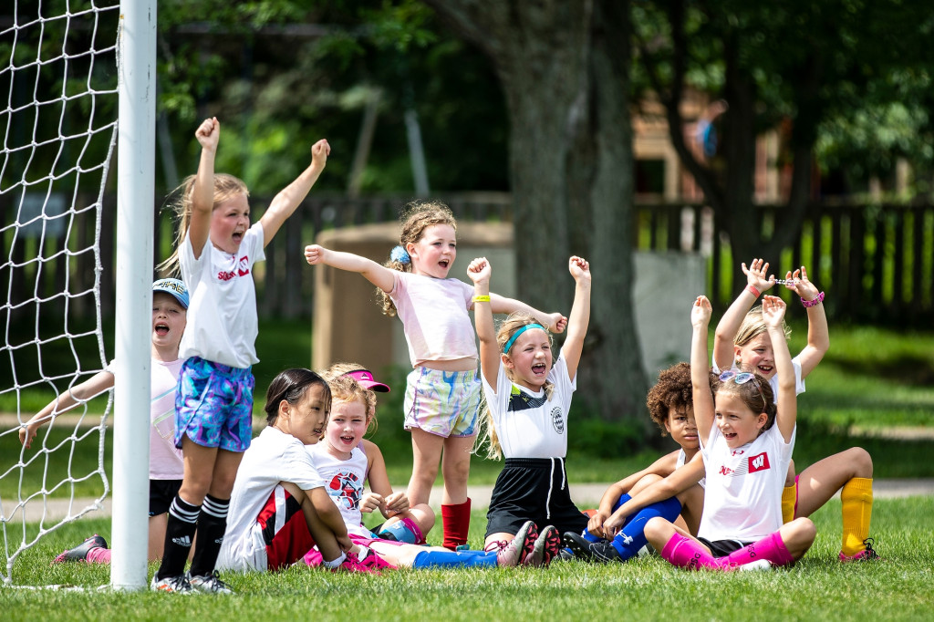A group of girls, about age 5, sit and stand near a soccer goal on an outdoor pitch. They are cheering and waving their arms in response to action not pictured.