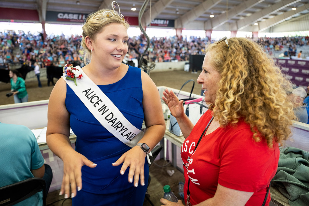 Taylor Schaefer wears her Alice in Dairyland tiara and sash and a blue sleeveless dress as she talks with Jennifer Mnookin (in a red Wisconsin T-shirt) in a livestock pavilion at State Fair.