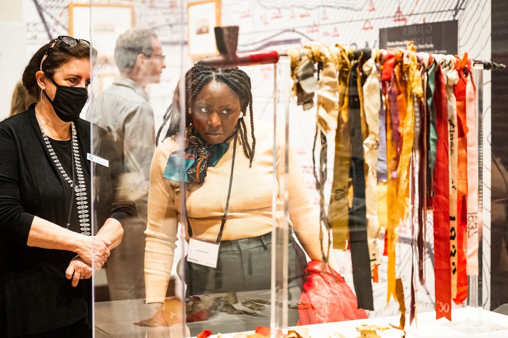 Two women look with skepticism at a display case holding a long smoking pipe with colorful ribbons tied along its stem.