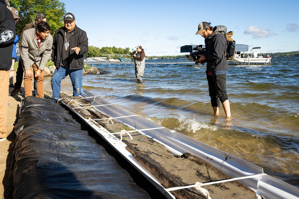 Casey Brown and Bill Quackenbush bend down to look at the remains of a canoe brought to the shore of Lake Mendota. A man with a video camera stands in the water filming them.