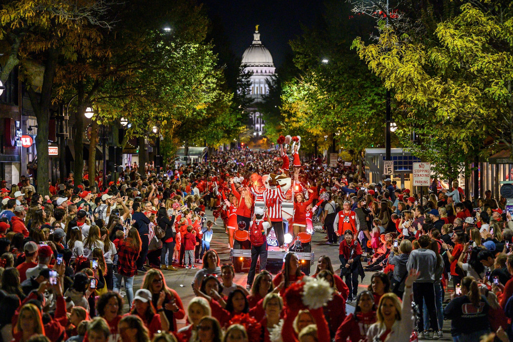 A wide shot at night of a homecoming float coming down State Street from the Capitol. Throngs of spectators surround the float carrying Bucky Badger and the UW Spirit Squad.
