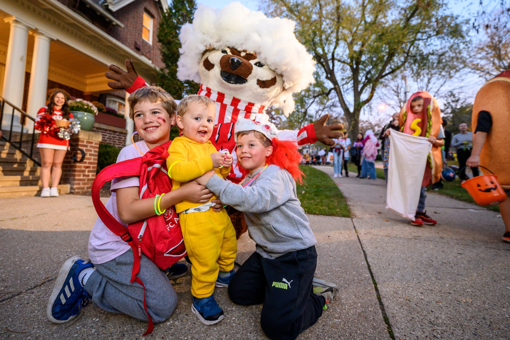 Three young children in Halloween costumes kneel on the ground and pose with Bucky Badger outside Olin House.