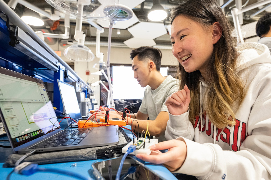 Two students sit at computers while holding circuit breakboards in their hands.