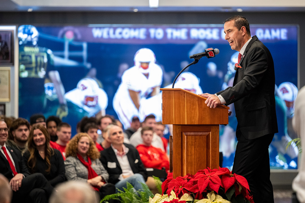 Luke Fickell stands in profile at a podium and speaks to a seated audience. A mural of UW football players is displayed behind him.