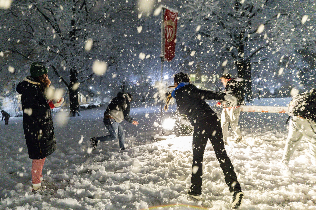 Five students in a circle pelt each other with snowballs as heavy snow falls at night on Bascom Hill.