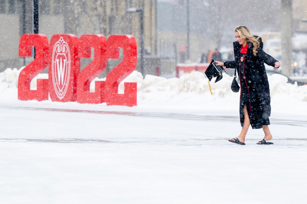 Wearing cap and gown and flip flops, Bella Persino holds her high heels in one hand as she walks up a very cold and snowy path to the Kohl Center. The a cutout of the year 2022 is on display in the background.