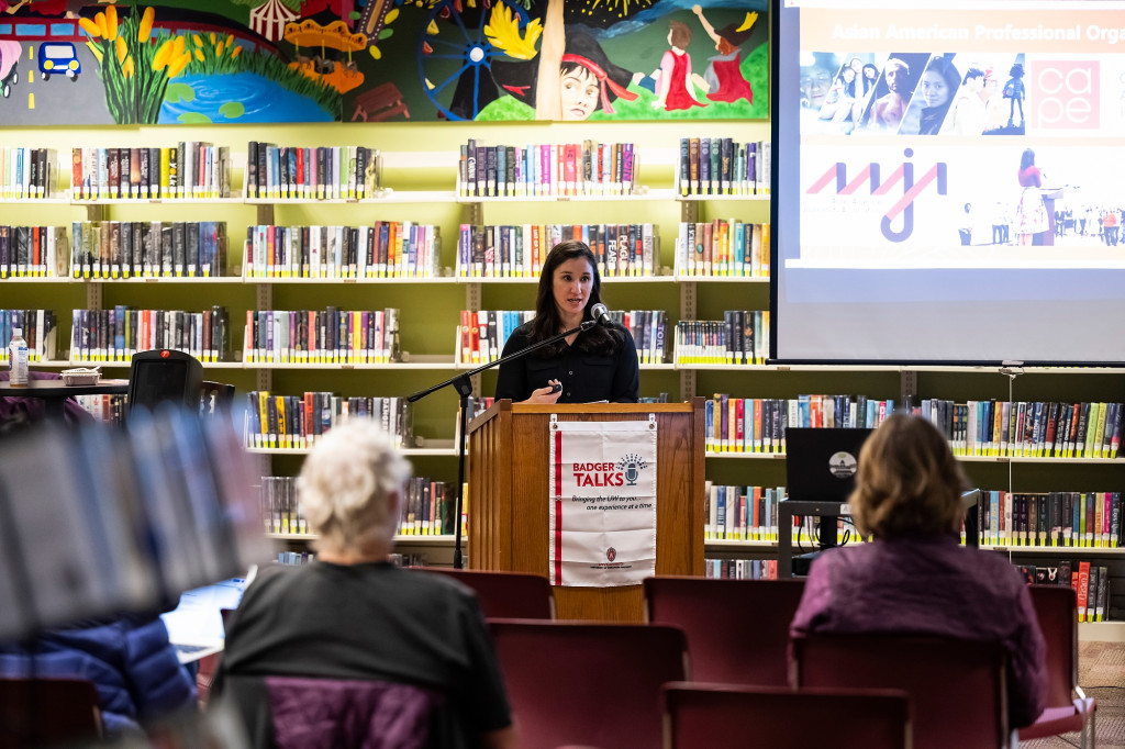 Lori Kido Lopez stands at a podium in a library setting. She stands in front of a shelf of books delivering a talk to a seated audience.