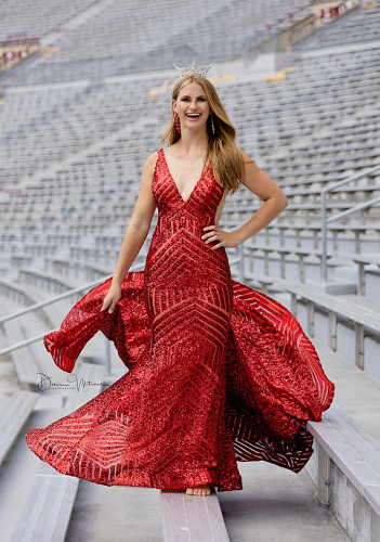 Portrait of Grace Stanke in a red ball gown and tiara standing on the bleachers at Camp Randall.