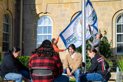 At back left, Sagen Quale and Aaron Bird Bear raise the Ho-Chunk Nation flag during a flag raising ceremony in honor of Indigenous Peoples Day in front of Bascom Hall. In the foreground, members of the Madtown Singers perform in a drum circle.